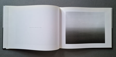 An open book, showing a minimalist interior spread with a large black-and-white landscape photograph on the right page and a mostly blank white left page with faint small text.