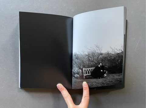 An open black-and-white photo book lies on a gray surface, with a hand holding the left page open. The right page shows a monochrome outdoor photograph of Nick and Susie on a bench, while the left page is solid black.