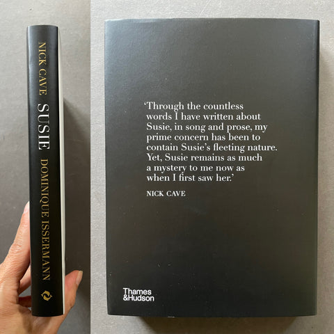 A hardcover book titled 'Susie' by Nick Cave and Dominique Issermann, shown standing upright next to a hand holding its spine. The front cover is matte black with white text containing a quote, and the spine is black with gold text.