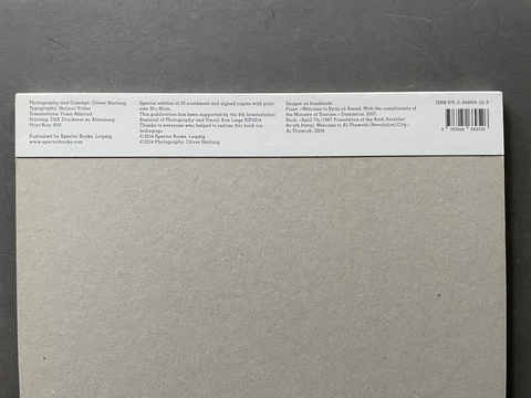 The back cover of a book or notebook, featuring a large textured gray cover with a white spine strip containing fine black text and a barcode, set against a dark gray background.