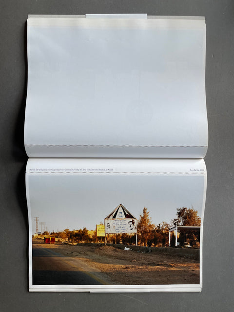An open glossy photo book or magazine lies flat against a dark gray background. The top page is blank white, while the bottom page features a landscape photograph of a rural road with a triangular warning sign, trees, and a small structure at sunset.