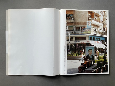 An open printed book or magazine laid flat on a dark gray surface, showing a mostly blank white upper page and a lower page with a colorful street-scene photograph and small text.