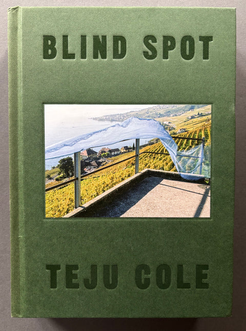 A hardcover book titled 'BLIND SPOT' by Teju Cole, featuring an olive green textured cover with a central photograph of a vineyard landscape under a white canopy, set against a dark gray background.