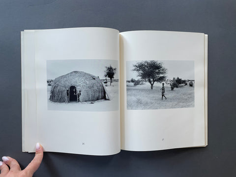 An open book or magazine spread against a dark gray background, showing two black-and-white landscape photographs printed on the pages: a thatched dome structure on the left page and a person walking outdoors on the right page. 