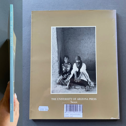 A hardcover book titled 'The African Desert' by Plossu, published by The University of Arizona Press, shown with its front cover and spine against a dark gray background. A hand is holding the left edge of the book.