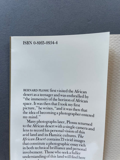 A close-up of a printed book or booklet page showing an ISBN number and a paragraph of text about Bernard Plossu and the African desert, photographed against a dark blue-gray surface.