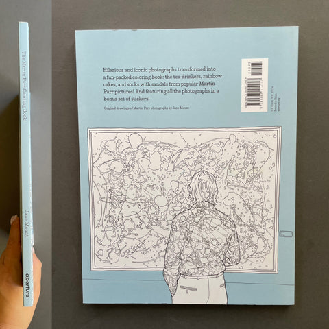 The back cover of the book is shown flat against a dark gray background, with a hand holding the spine on the left. The cover is light blue with black text and a black-and-white line art illustration of Martin Parr.
