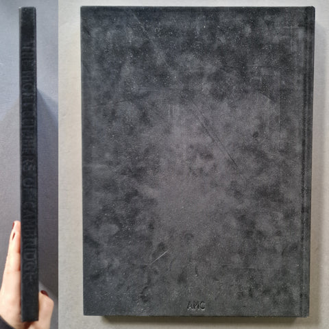 A hardcover book shown from the front and side, with a hand holding the spine. The cover is dark charcoal gray with a mottled texture and the embossed brand name AMC at the bottom.