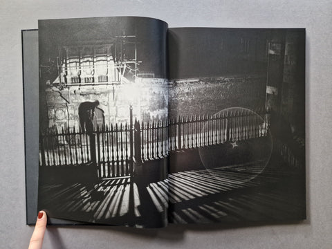 An open hardcover book displaying a high-contrast black and white photographic print of a person standing behind a fence in a moody urban setting.