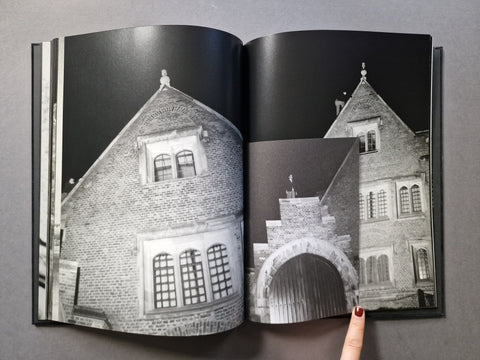 An open hardcover book showing black-and-white architectural photographs of brick buildings and a stone archway, with a finger pointing at the right page, on a gray surface.