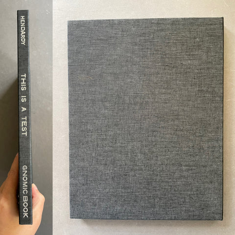 A hardcover book with a textured gray linen cover and white text on the spine, shown next to a hand holding the spine. The front cover is plain and the spine reads 'HENDARDY THIS IS A TEST GNOMIC BOOK'.