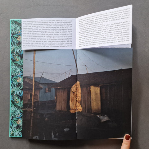 An open book, showing printed text on the inner pages and a large photograph of rustic wooden huts with a yellow garment hanging from a pole. A hand with dark red nail polish is visible at the bottom right edge.