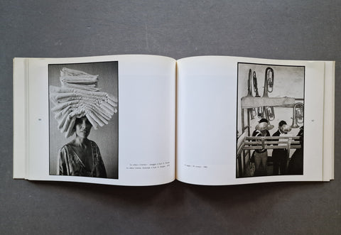An open book laid flat on a dark gray surface, showing two black-and-white printed photographs on the pages. The left page contains a tall portrait of a person balancing a stack of sheets on their head, and the right page shows a black-and-white interior scene with several people and trumpets.
