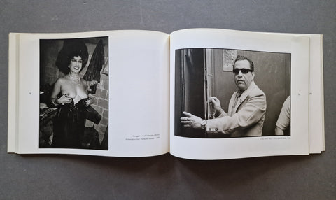 An open book laid flat on a dark gray surface, showing two black-and-white printed photographs on the pages - one of what appears to be a semi naked female performer and one of a man with dark sunglasses.