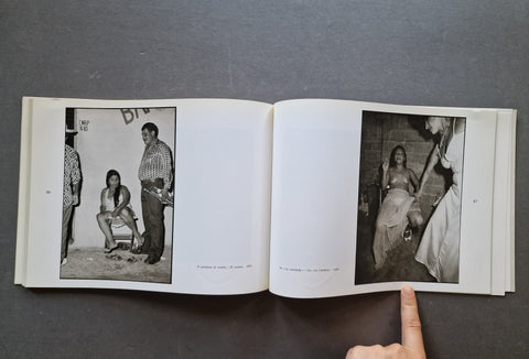 An open book laid flat on a dark gray surface, showing two black-and-white printed photographs on the pages - both photos depict women sitting on chairs - one is semi naked and smoking a cigarette.
