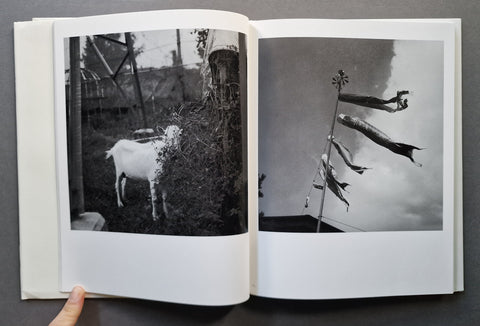 An open book showing two black-and-white photographic prints on the pages, photographed against a dark gray background. A human finger is visible holding the left edge of the book.
