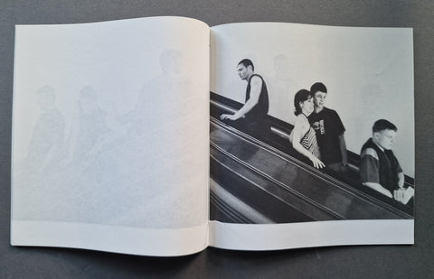 An open book, showing a black-and-white printed photograph of several people riding an escalator on the right page and a faint large monogram or logo on the left page.