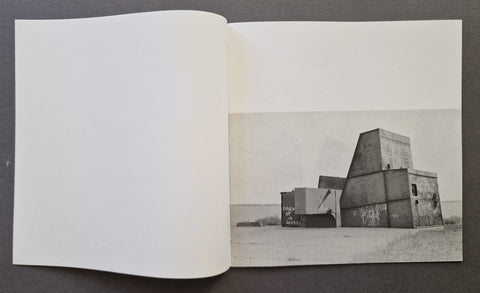 An open book, showing a black-and-white architectural photograph printed on the right page. The left page is blank, and the photo depicts a large concrete structure in an open desert landscape.