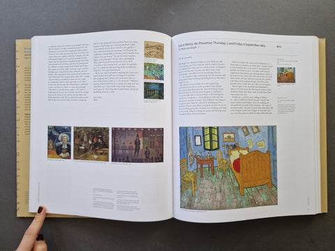 An open book spread showing printed text and multiple paintings, photographed from above on a dark gray surface. A hand with dark nail polish is visible holding the left page edge.