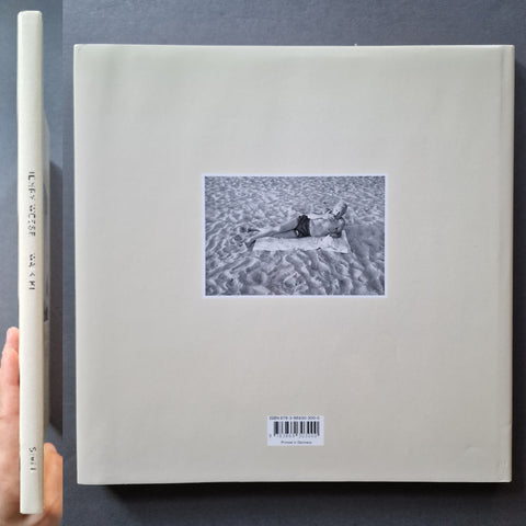 A square art book, featuring a central black-and-white photograph of a person lying on sand. 