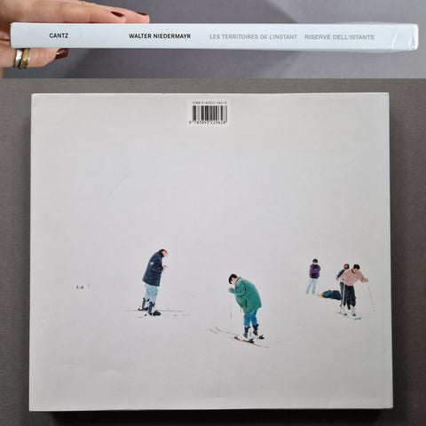 A hand holds a white book or album cover at the top edge of a larger square white cover below. The lower cover has a minimalist illustration of several skiers on a snowy field.