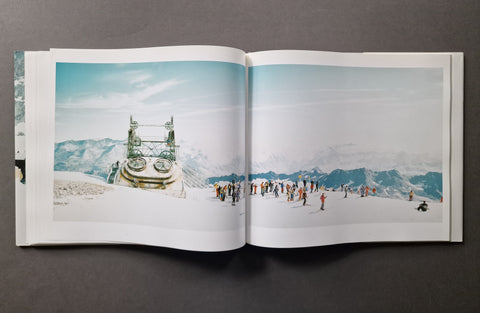 An open hardcover photo book , showing a snowy mountain landscape printed across both pages. The scene includes a large metal structure on the left page and small people on the snowy mountain on the right page.