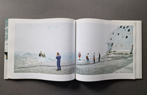 An open hardcover book, showing a large printed photograph spread across both pages. The photo depicts a snowy mountain landscape with several small people standing near a white building.