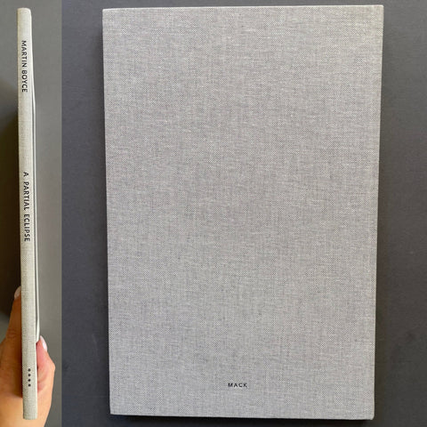 A hardcover book titled 'A Partial Eclipse' by Martin Boyce, featuring a light gray linen-textured cover and spine, held next to the book by a hand against a dark gray background.