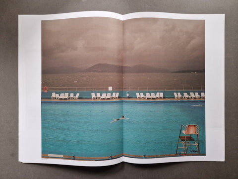 An open publication spread photographed from above on a dark gray surface, featuring a large photographic print of a swimming pool with lounge chairs, distant hills, and a cloudy sky.