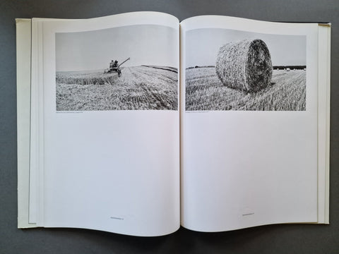 An open book spread showing two black-and-white photographs of a field scene: a combine harvesting on the left page and a large hay bale on the right page, photographed against a dark gray background.