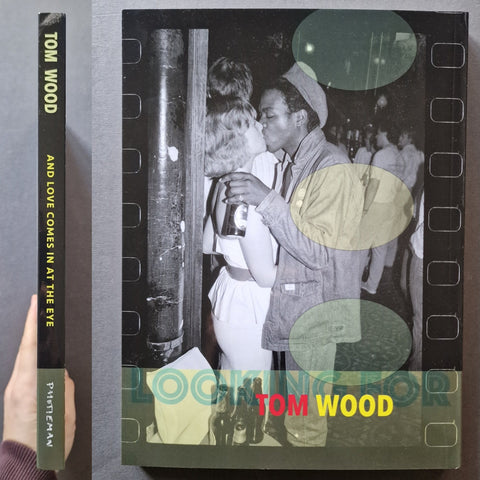 A book titled "Tom Wood: And Love Comes In At the Eye" shown standing upright with its spine visible, photographed against a dark gray background. A hand is holding the left edge of the book.