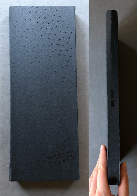 A dark gray rectangular sharpening stone with a textured surface and small perforations, shown next to its slim black spine on a gray background. A hand is holding the spine to show its thickness.