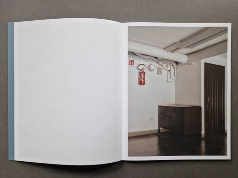 An open book, showing a large blank white page on the left and a printed interior scene on the right with a room, exposed ceiling pipes, a red fire extinguisher, and a dark cabinet.