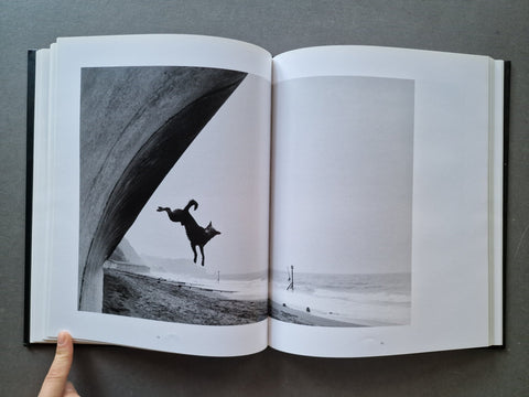 An open hardcover book showing a black-and-white photographic print of a dog leaping under a large arch near a beach. 