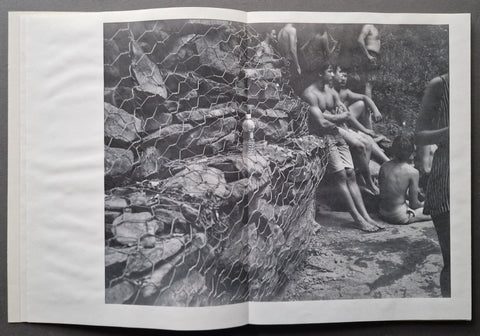 An open book spread showing a black-and-white photograph of several shirtless men sitting and standing near a rocky wall with wire mesh in the foreground.
