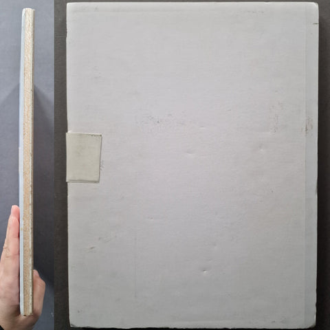 A large square off-white panel or board shown from the back, with a hand holding a slim side profile view beside it. The back has a small rectangular tab or fastener and a slightly rough, matte texture.