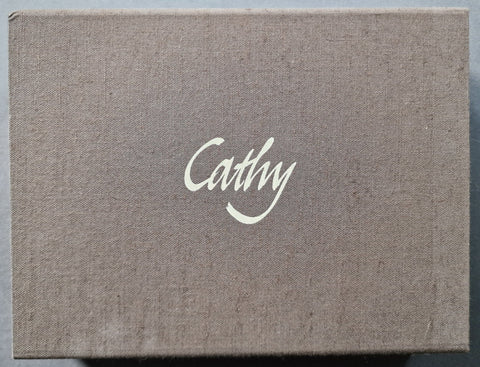 A square fabric-covered box with a textured taupe-brown linen-like surface and the name 'Cathy' printed in white script at the center.