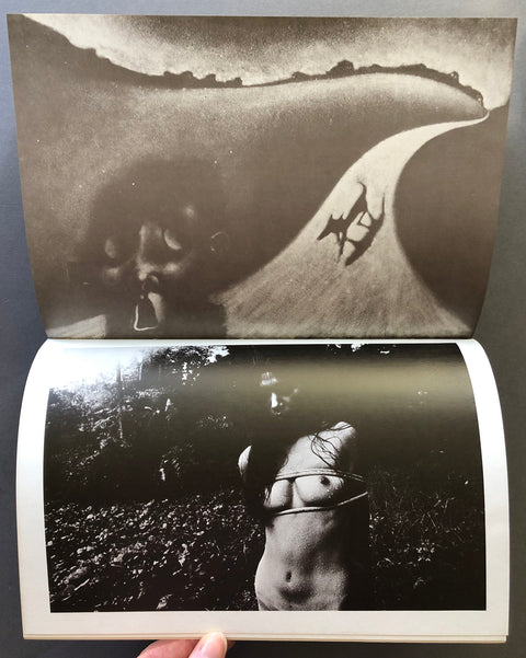 An open book or photo album showing two black-and-white photographic prints. The top page displays a surreal landscape-like scene with a curved path and a dark face in the background, while the bottom page shows a dark, moody photograph of a nude torso in a forest.