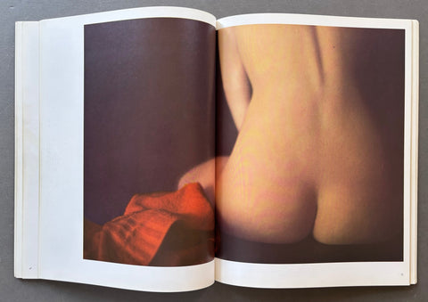 An open book or magazine spread showing a large photographic print of a nude female back with warm skin tones against a dark background, with a red fabric element in the lower left of the photo.