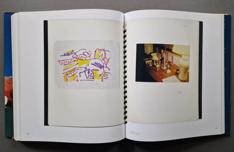 An open hardcover book, showing two interior pages with black binding strips. The left page contains a small abstract artwork in purple, yellow, and orange lines on a white sheet, and the right page contains a small color photograph of a candlelit table scene.