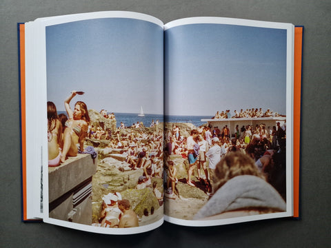An open hardcover book spread, showing two printed pages with a large vintage beach photograph. The photo depicts a crowded beach scene with people, rocks, blue sky, and ocean.