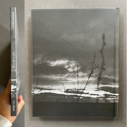 A hardcover book or art print with a black-and-white landscape photograph on the cover, shown standing upright next to its spine. A hand is holding the spine at the left edge.