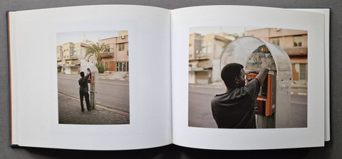 An open hardcover book. The left page contains a sepia-toned photo of a person standing near a street building, and the right page shows a color photo of a person interacting with a metallic vending or coin machine in an outdoor urban setting.