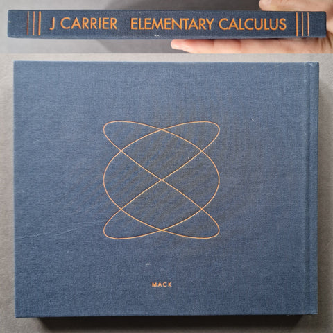 A hardcover textbook titled 'J Carrier Elementary Calculus' by Mack, shown closed with a hand holding the top edge of the spine above it. The cover is textured slate blue with a gold geometric orbit design in the center and gold lettering.