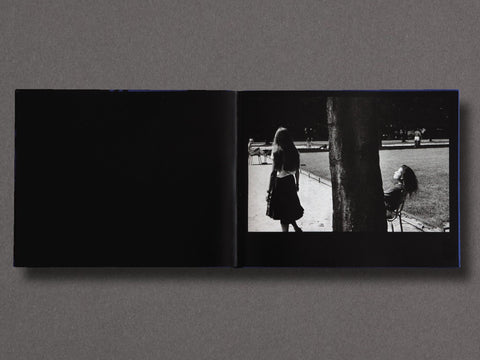 An open black book laid flat on a gray surface, showing a black-and-white photograph printed on the right page with a silhouetted figure walking beside a tree in an outdoor park setting.