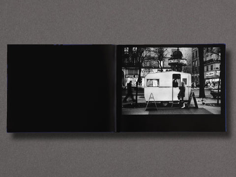 An open black-and-white photo book lies flat against a dark gray background. The left page is solid black, while the right page shows a black-and-white street photograph of a small trailer or food cart with pedestrians and storefronts in the background.