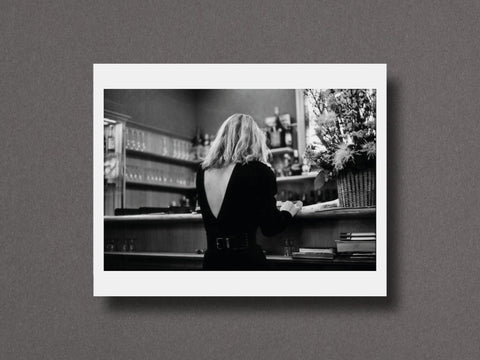 A black and white photographic art print featuring a woman seen from behind at a bar, displayed with a wide white border against a textured gray background.