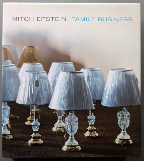 Mitch Epstein Family Business book cover featuring a photograph of multiple table lamps with light blue pleated shades and clear glass bases on a dark surface against a pale background.