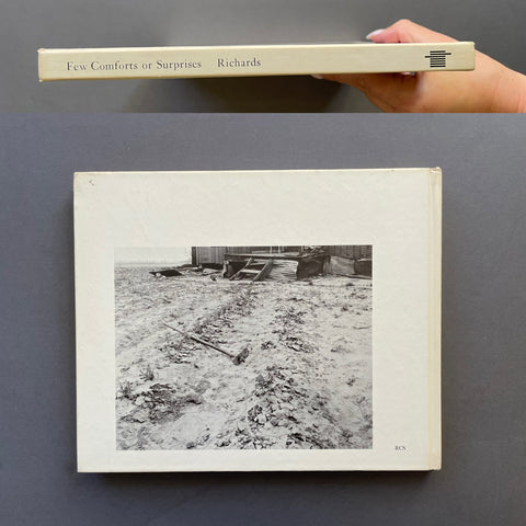 A hardcover art book titled 'Few Comforts or Surprises' by Eugene Richards is shown, with the top edge held by a hand at the top of the frame. The book has a cream-colored cover featuring a black-and-white photograph of a snowy, debris-strewn outdoor area.