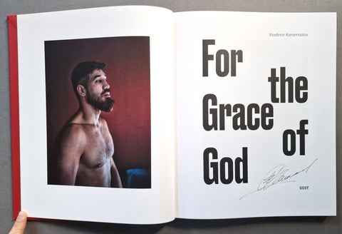 An open book laid flat on a gray surface, showing a printed page with a portrait photo on the left and large black text reading 'For the Grace of God' and a handwritten signature on the right. 